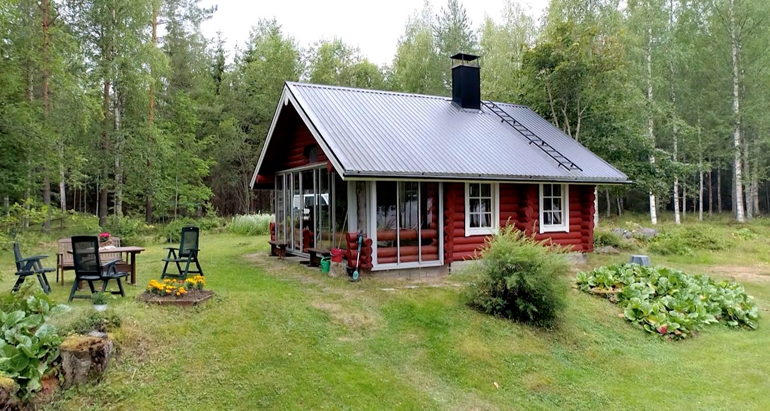 Punainen pihamökki harmaalla katolla metsän laidassa, nurmella tuolit, pöytä ja kukkapenkki