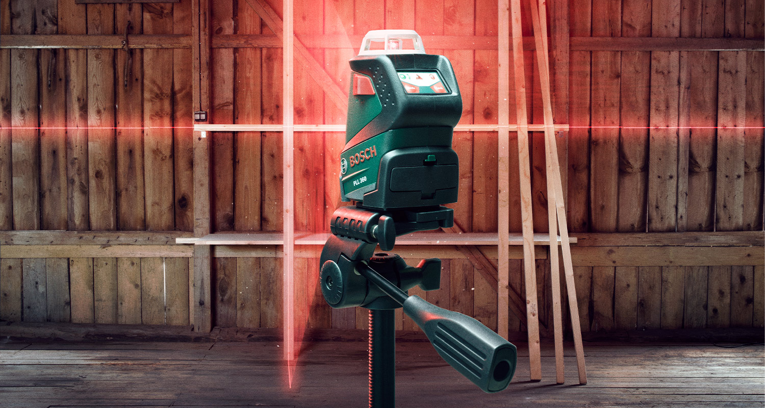 Vihreä Bosch-laserasema jalustalla heittää punaiset vaak- ja pystyviivat puuseinälle.