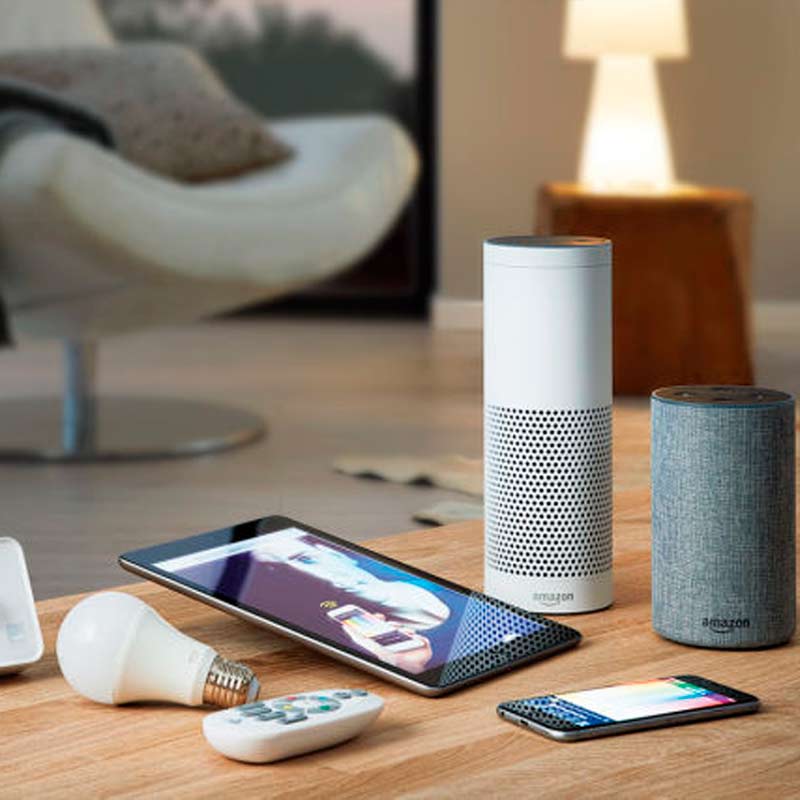 Olohuoneen pöydällä valkoinen ja harmaa Amazon Echo -kaiutin, tabletti, puhelin ja lamppu.