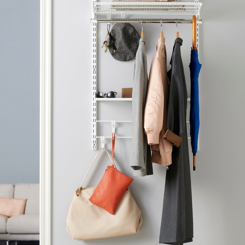 Seinänaulakossa harmaa ja beige takki, oranssi pussukka, sininen sateenvarjo vaaleaa taustaa vasten