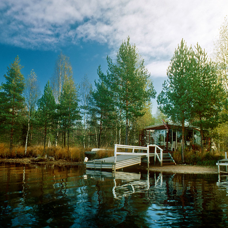 Punainen lasiseinäinen rantasauna mäntyjen reunustamalla järvenlaiturilla.