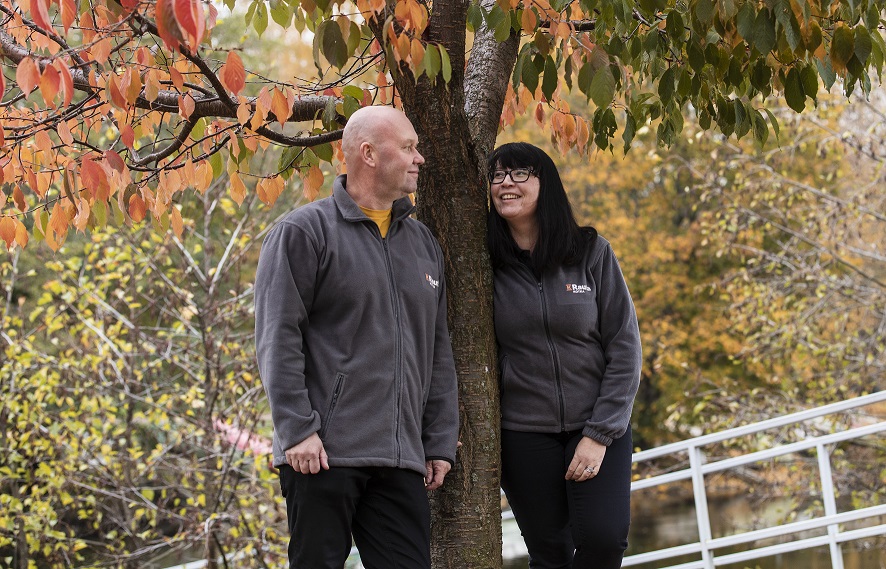 Kaksi harmaatakkista ihmistä puun luona, taustalla vesistö ja syksyn oranssi-keltaiset lehdet.