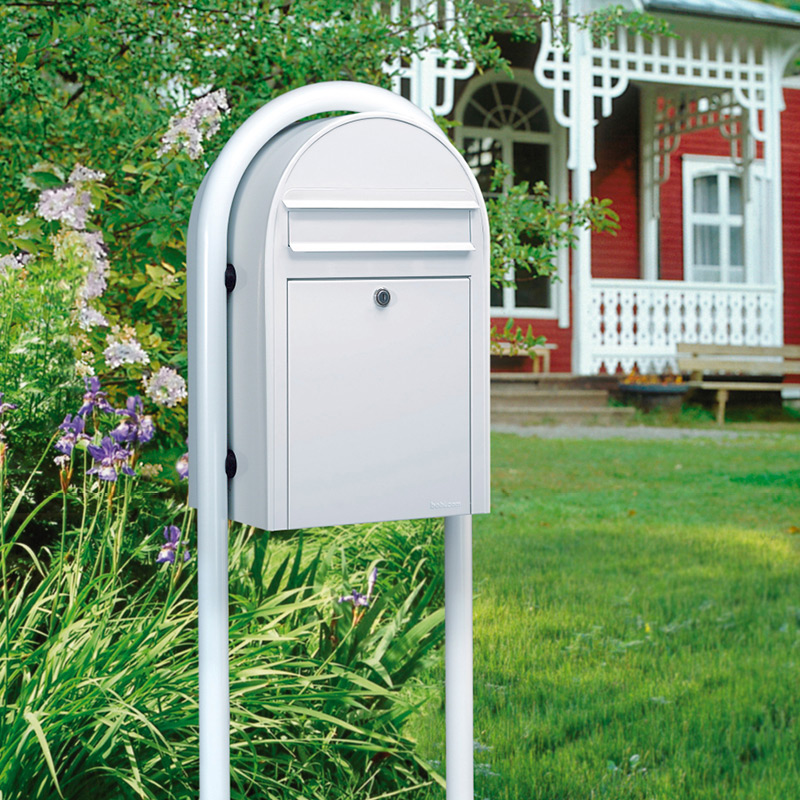 Valkoinen postilaatikko jalustassa nurmikolla, taustalla punavalkoinen puutalo ja kurjenmiekkoja