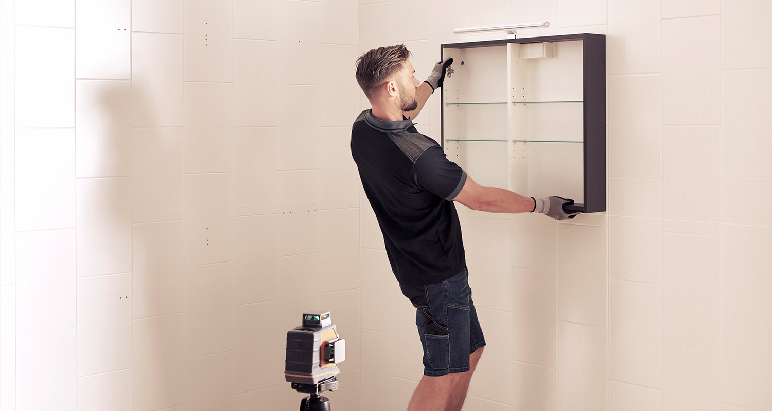 Asentaja kiinnittää mustavalkoista peilikaappia valkoiselle laatoitetulle seinälle.