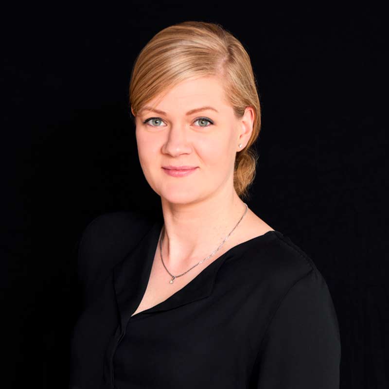 Vaaleahiuksinen henkilö mustassa paidassa ja hopeisella kaulakorulla tummalla taustalla.