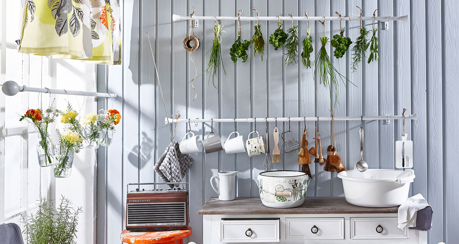 Keittiössä vaaleansinisen lautaseinän edessä roikkuvat vihreät yrtit, kukat ja valkoiset mukit.