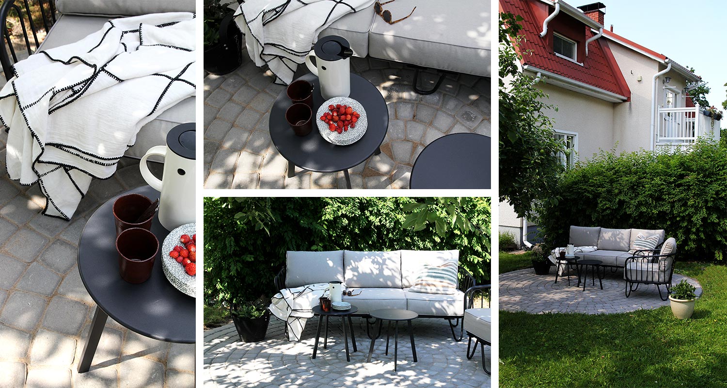 Ulkoterassilla harmaa sohva ja nojatuoli, mustat pyöreät pöydät, valkoinen viltti ja mansikoita.