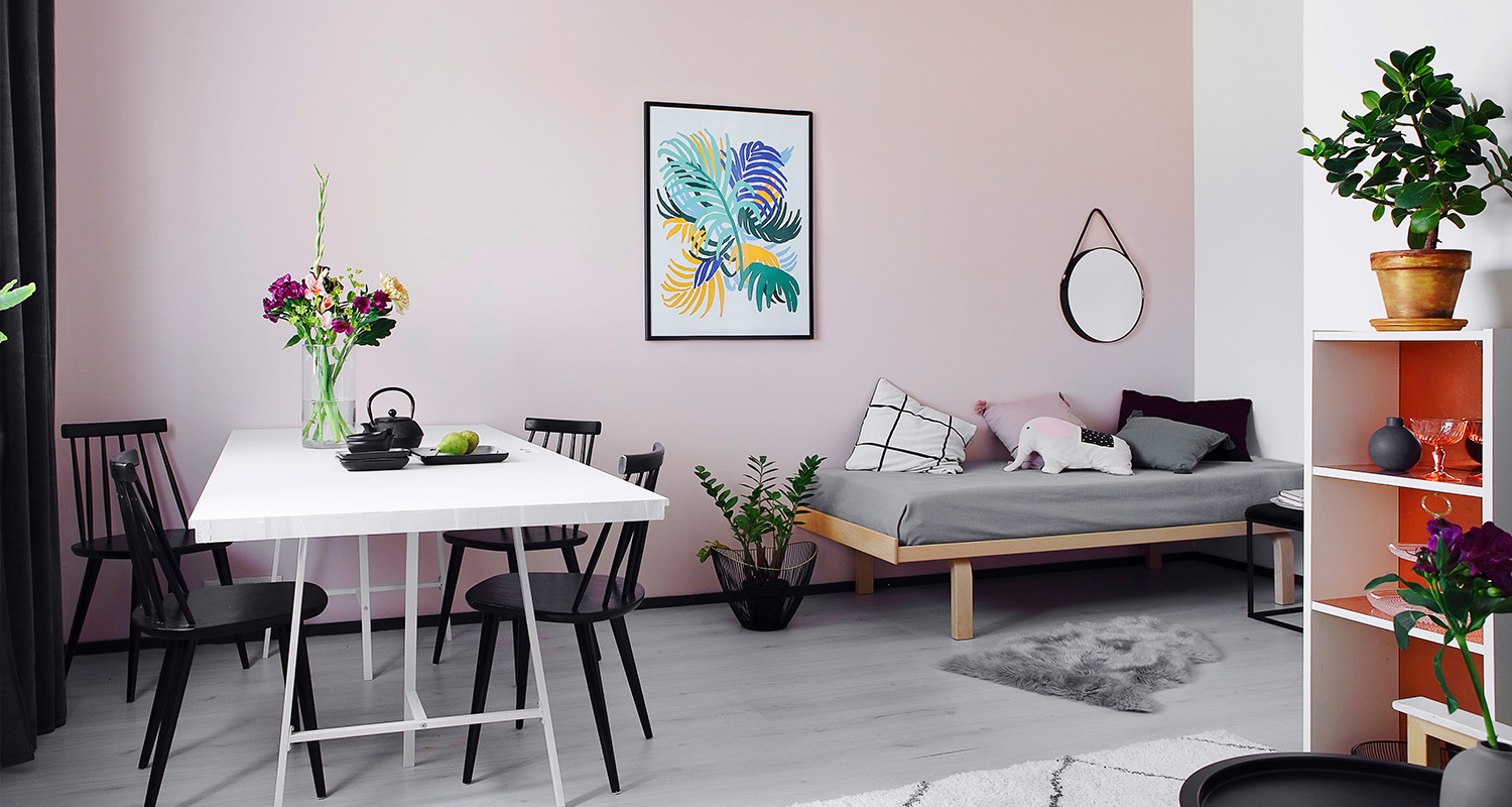 Vaaleanpunainen huone: valkoinen pöytä mustilla tuoleilla, kukkakimppu ja harmaa vuodesohva.