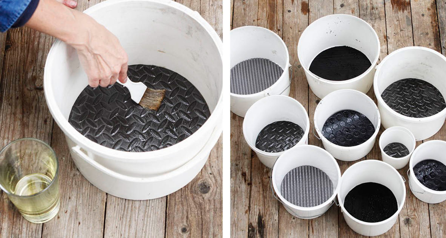 Käsi siveltimellä maalaa mustaa kumimattoa valkoisessa ämpärissä, puulattialla muita ämpäreitä