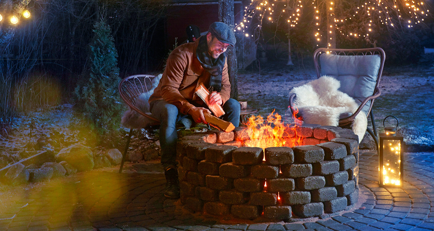 Mies lisää halkoja tiiliseen nuotioon lumisella pihalla, ympärillä vilkkuvat valosarjat.