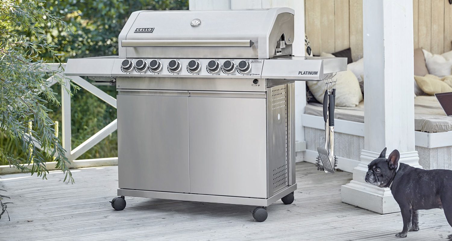 Hopeanvärinen kaasugrilli terassilla, grillivälineet roikkuu, etualalla musta bulldoggi.