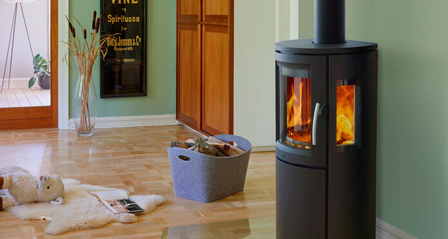 Mustapyöreä puu-uuni palaa vaaleanvihreän seinän edessä, koivuparketilla huopakori polttopuineen.
