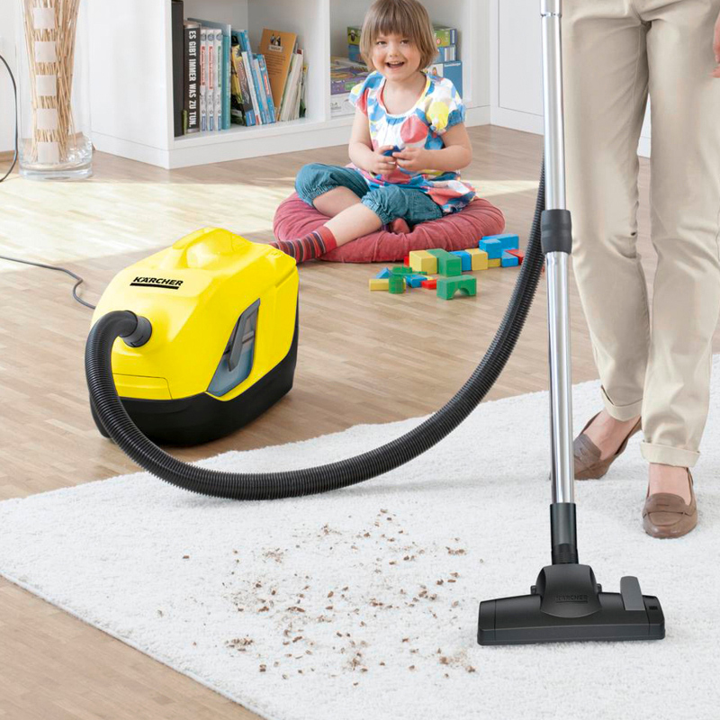 Keltainen pölynimuri ja imuroiva aikuinen valkoisella matolla, lapsi leikkii taustalla.
