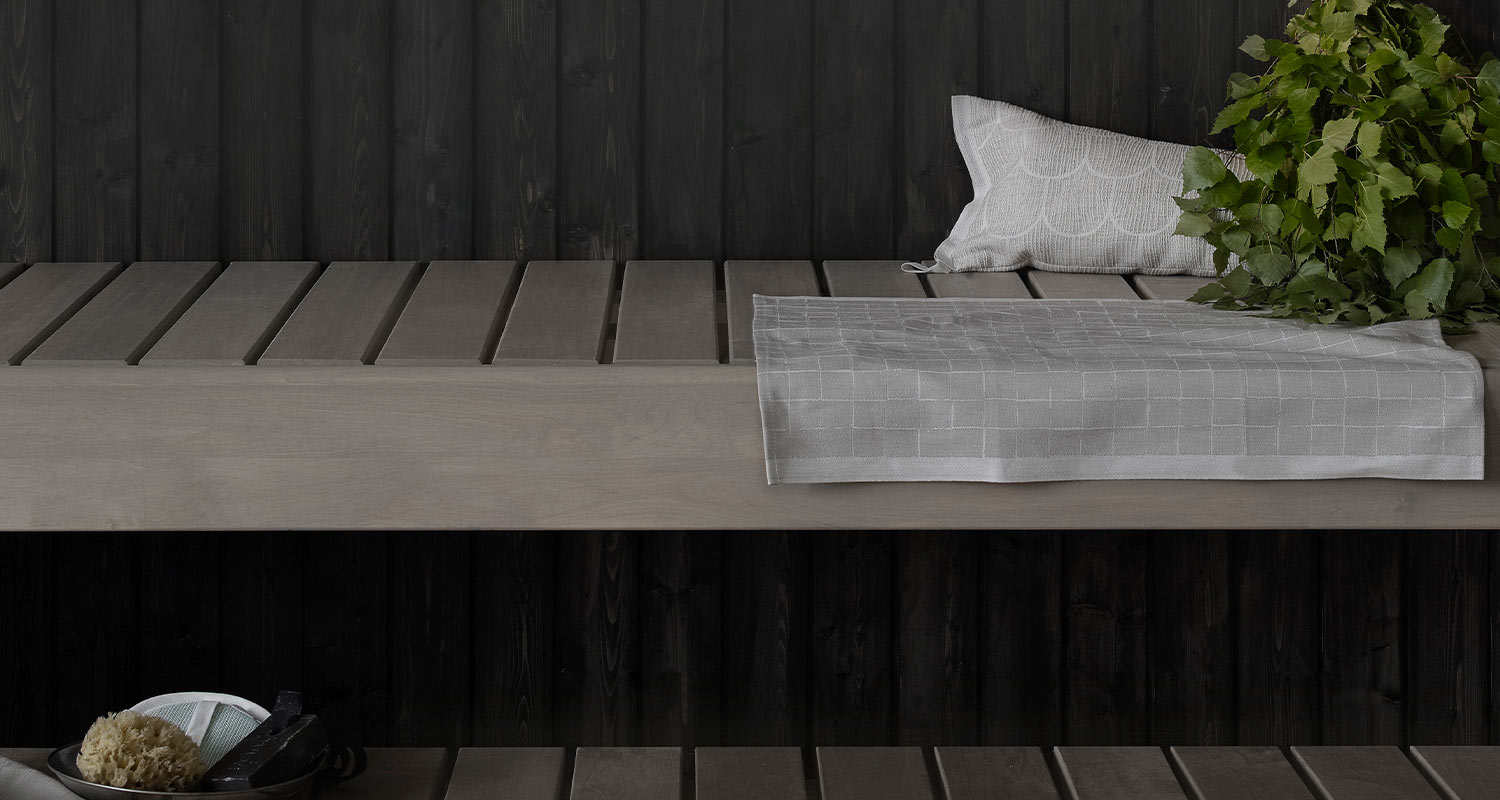 Vaalea puupenkki tumman lauta­­seinän edessä, päällä harmaa liina, tyyny ja vihdat.