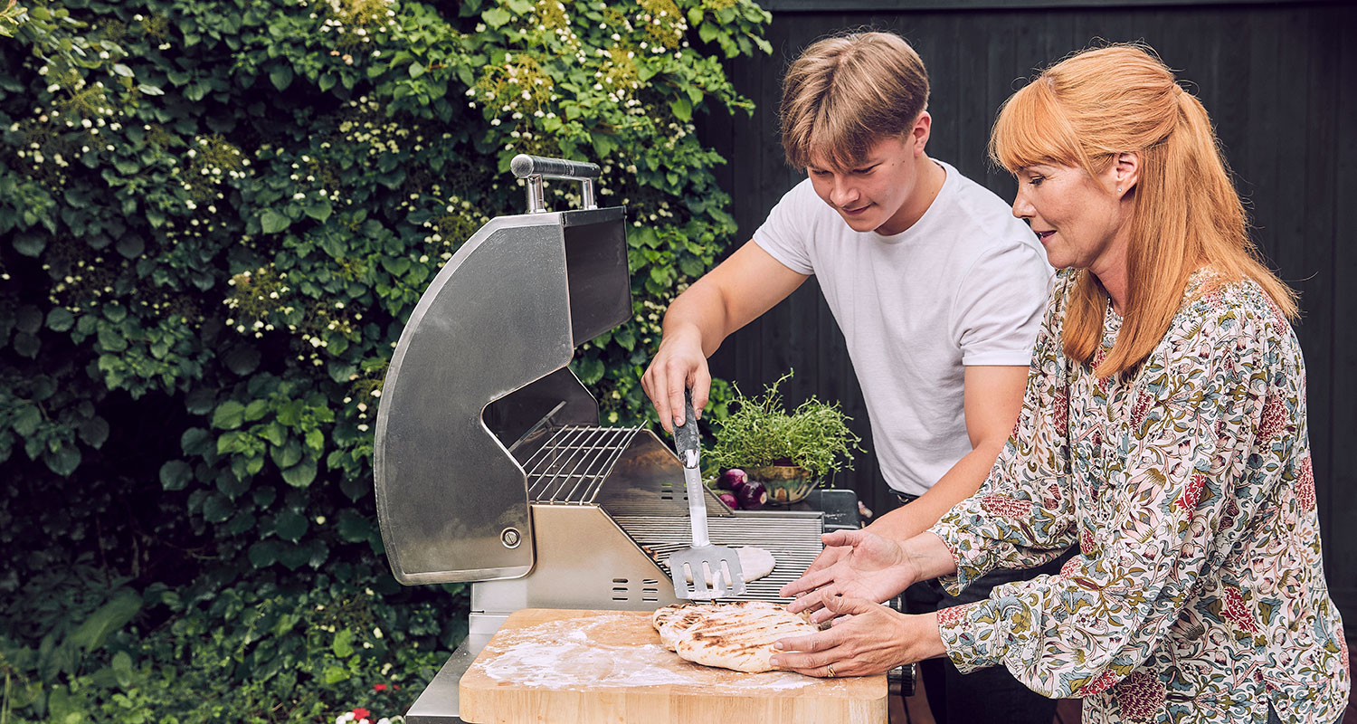 Mies paistaa pizzapohjia kaasugrillissä, nainen muotoilee taikinaa leikkuulaudalla viheraidan edessä