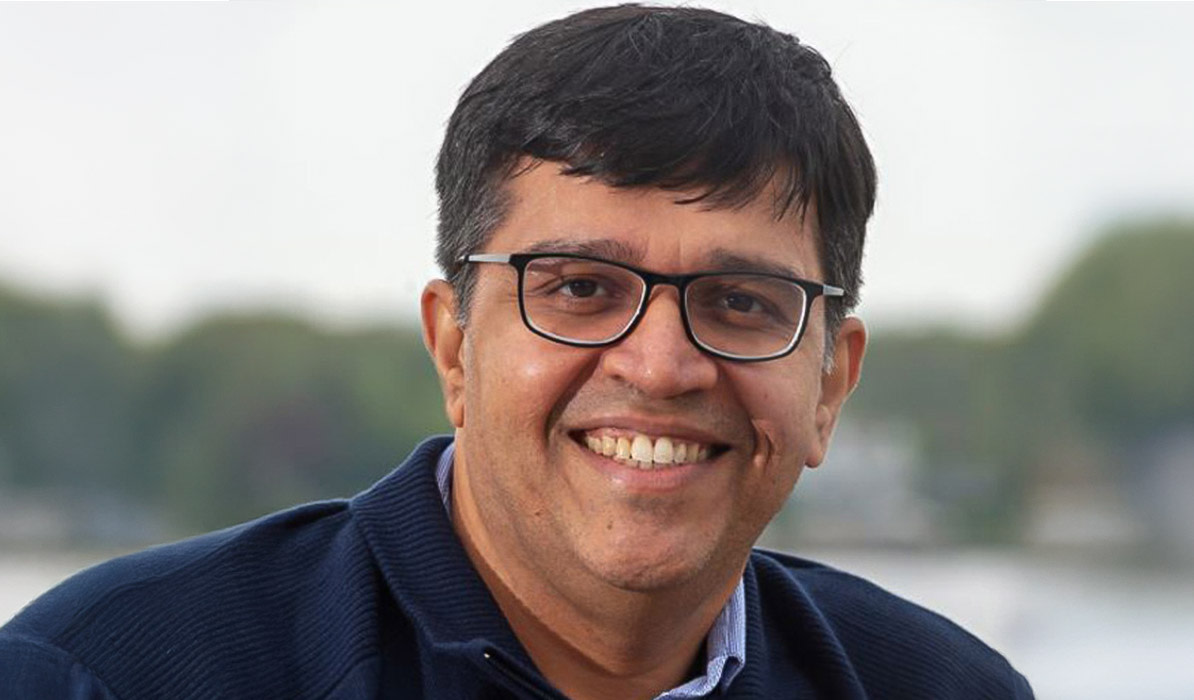 Man with glasses wearing a navy blazer, smiling outdoors with water in the background.