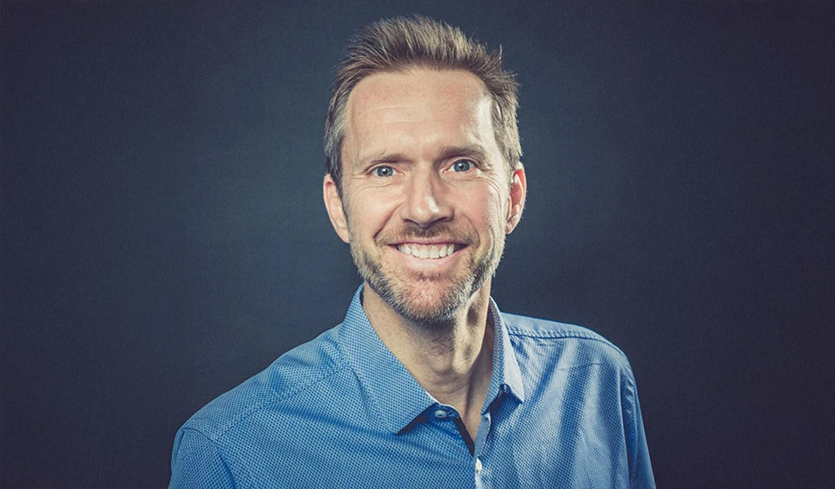Smiling man with short hair, wearing a blue collared shirt, posing in front of a dark background.
