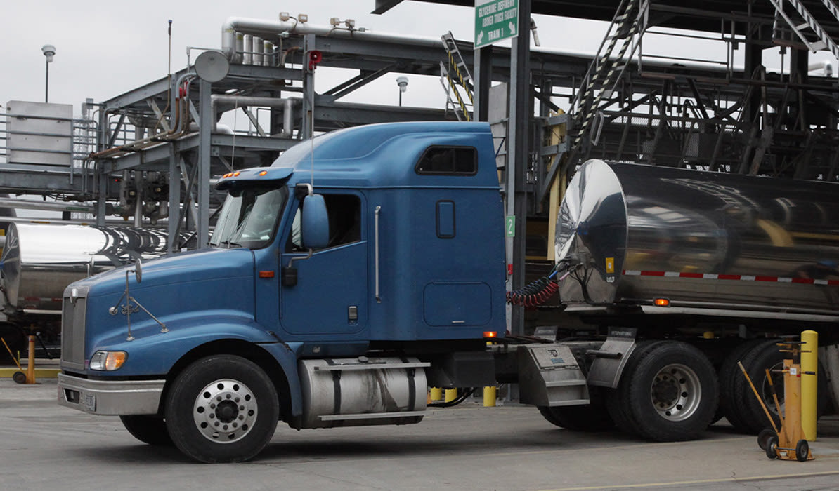 New blue semi-truck with a tanker trailer parked at a loading facility in an industrial area on a cloudy day.