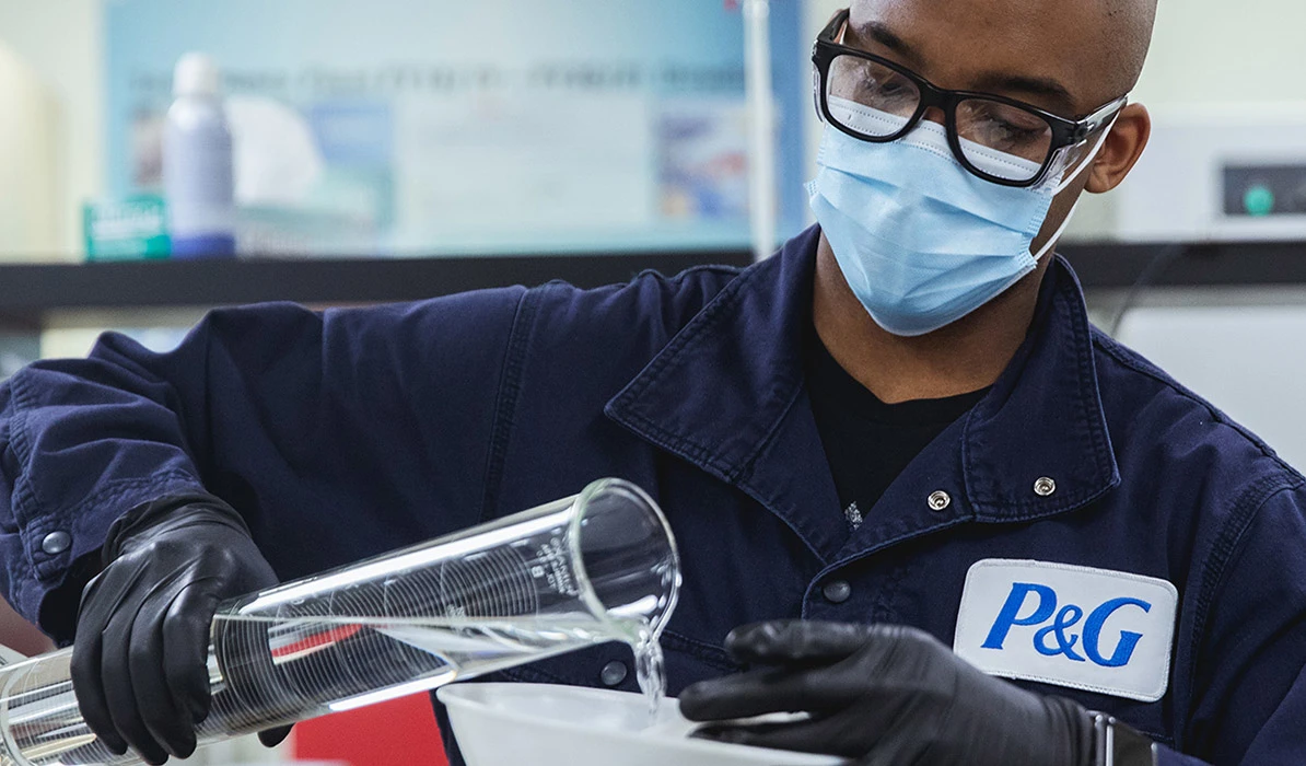 A masked and gloved technician wearing a P&G uniform carefully pouring liquid into a large glass container in a lab setting.