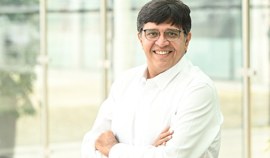 Smiling man wearing glasses and a white shirt standing with arms crossed in front of large windows