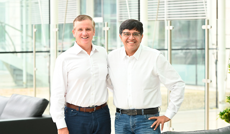 Two men standing side by side indoors, both wearing white shirts and smiling at the camera.