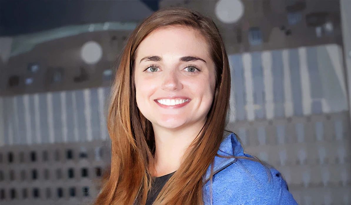 Smiling woman with long brown hair, wearing a blue blazer, posing in front of a blurred background.
