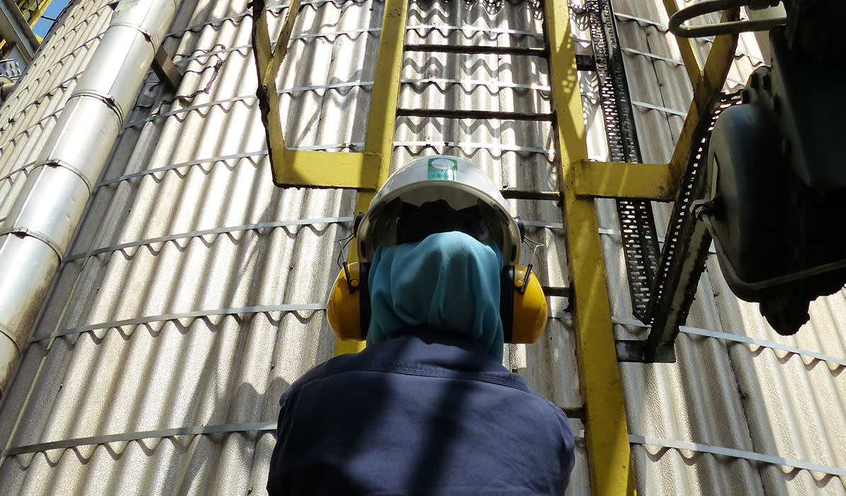 Industrial worker wearing a safety helmet, ear protection, and a blue uniform, climbing a yellow ladder on a metal silo structure.