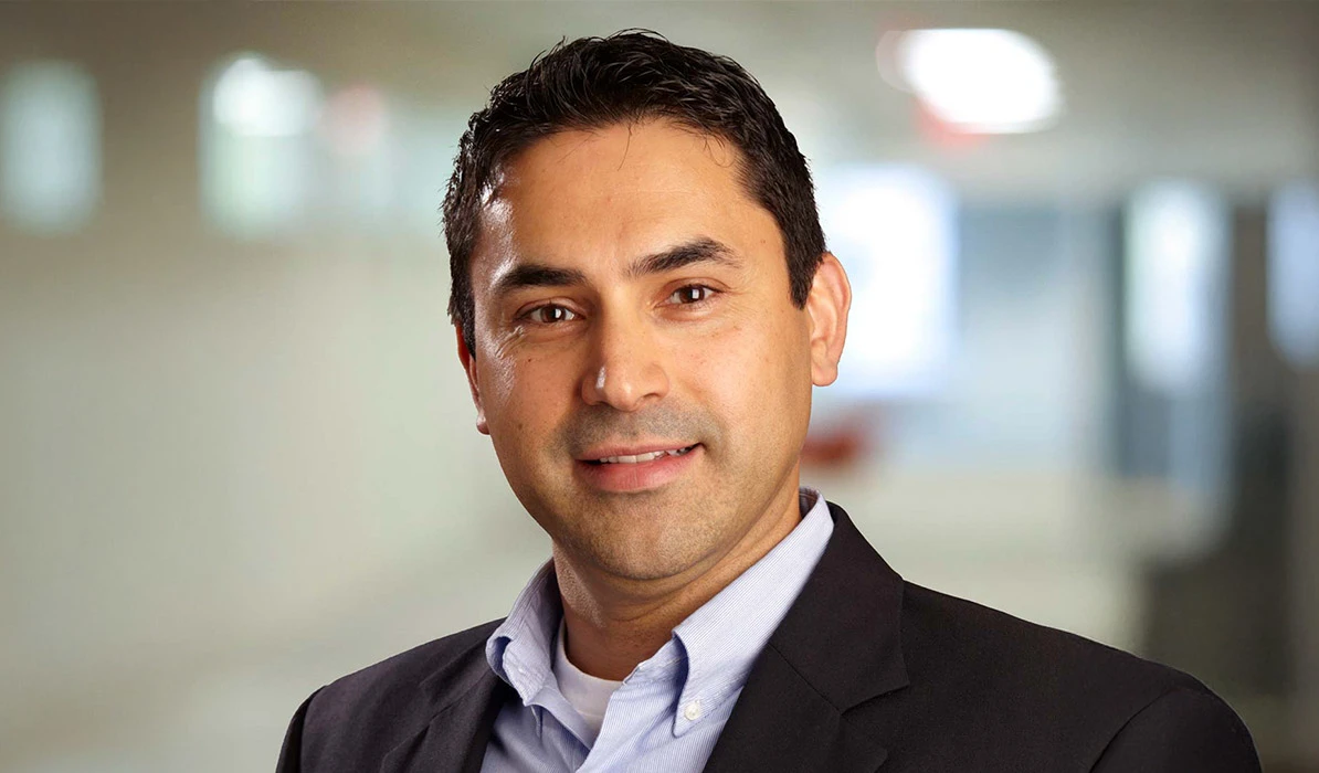Smiling man with short dark hair, wearing a black blazer and light blue shirt, posing indoors with a blurred office background.