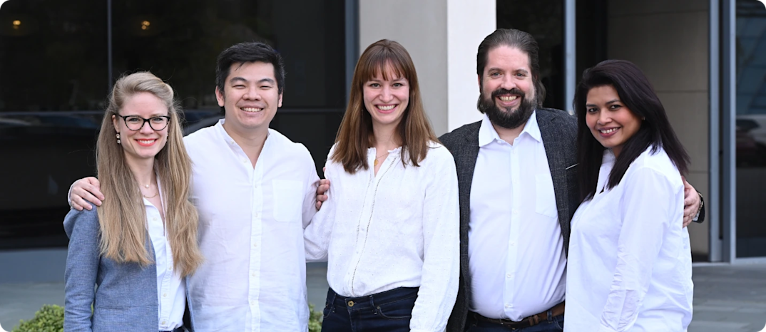 Five colleagues in business-casual attire standing side by side with arms around each other, smiling at the camera in front of an office building.