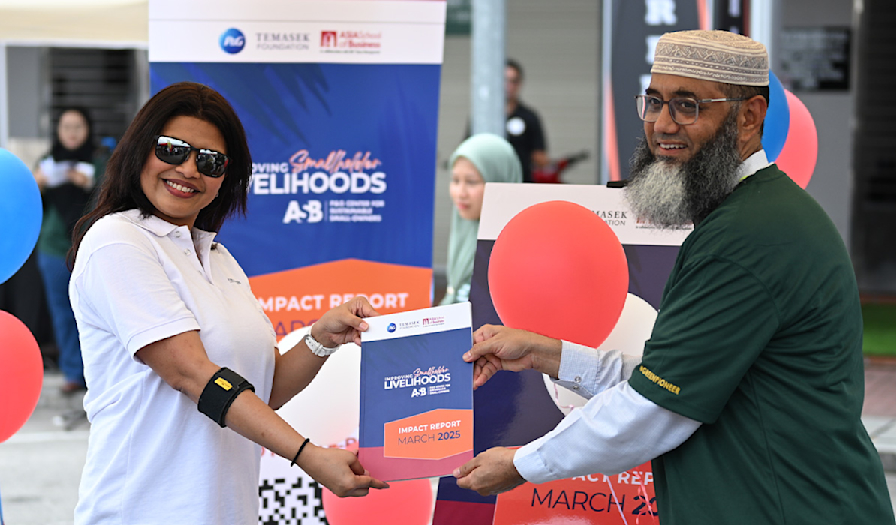 A woman in a white shirt and sunglasses presenting a certificate to a man in a green shirt and cap during a community event, with balloons and banners in the background.