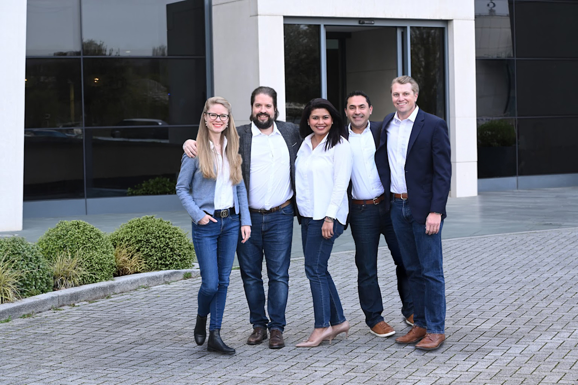 Five colleagues in business-casual attire standing side by side with arms around each other, smiling at the camera in front of an office building.