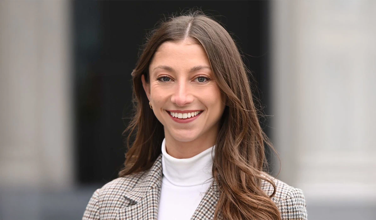 Smiling woman with long brown hair, wearing a plaid blazer and white turtleneck, posing outdoors with a blurred building in the background.