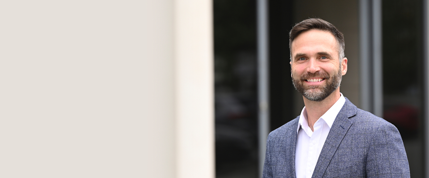 A smiling, bearded man wearing a white collared shirt and a gray blazer, standing in front of a blurred outdoor background.