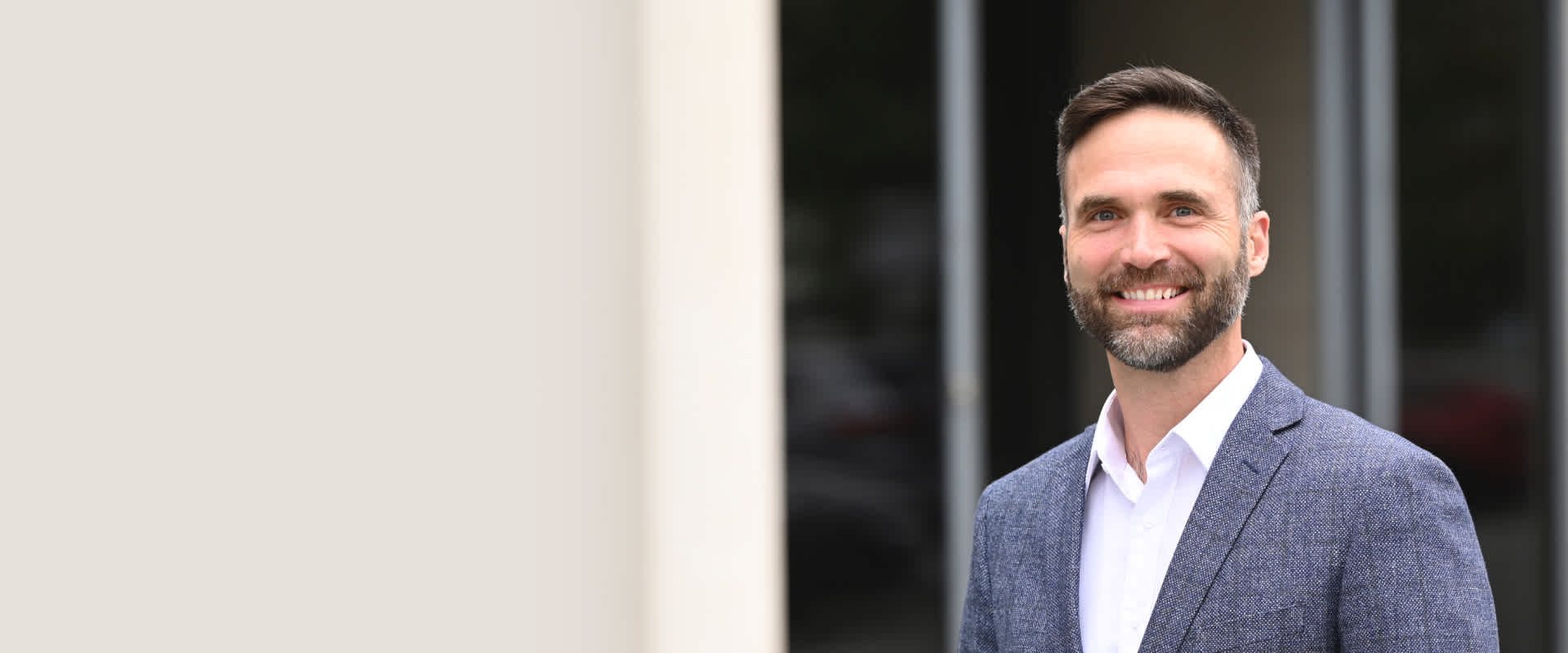 A smiling, bearded man wearing a white collared shirt and a gray blazer, standing in front of a blurred outdoor background.