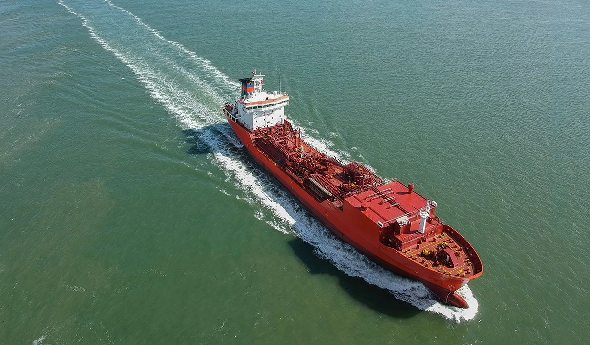 A red cargo ship navigating through calm waters, leaving a wake behind as it sails across the sea under bright sunlight.