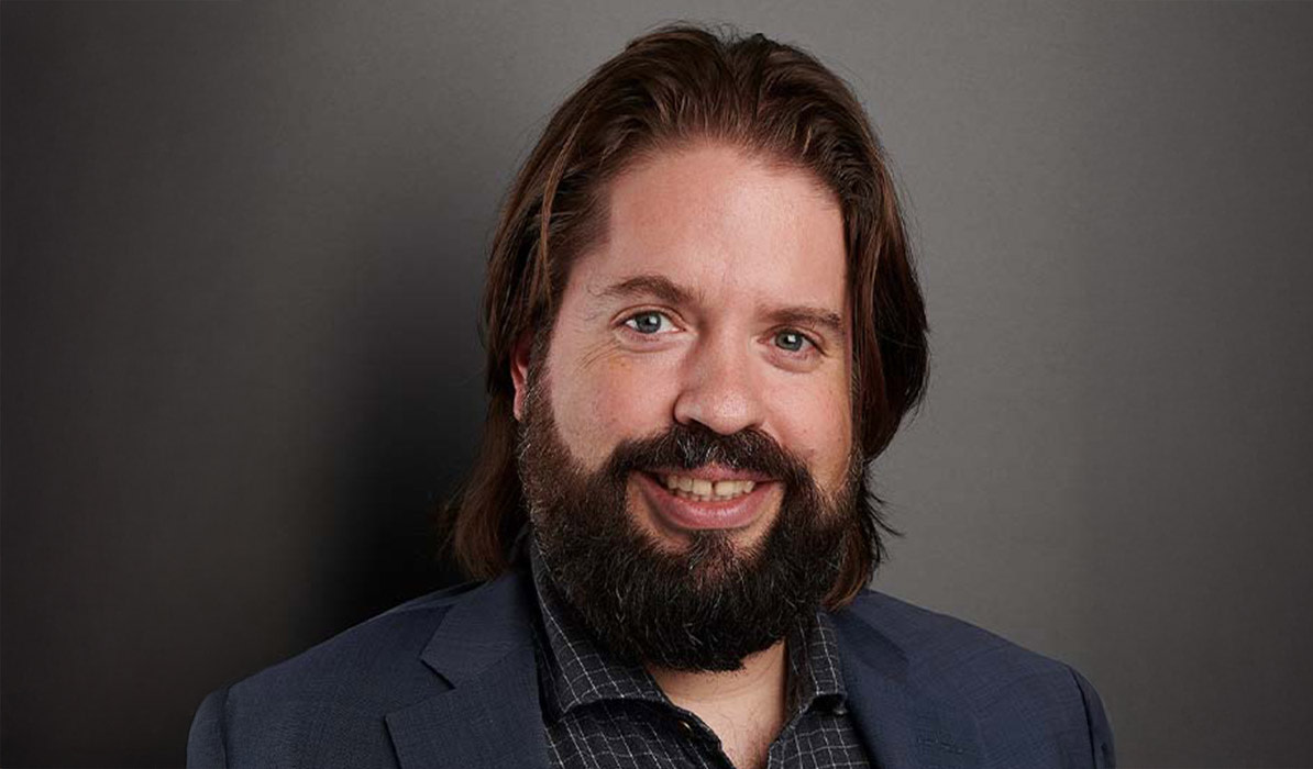  Smiling man with long hair and a beard, wearing a dark suit and checkered shirt, posing in front of a gray background.