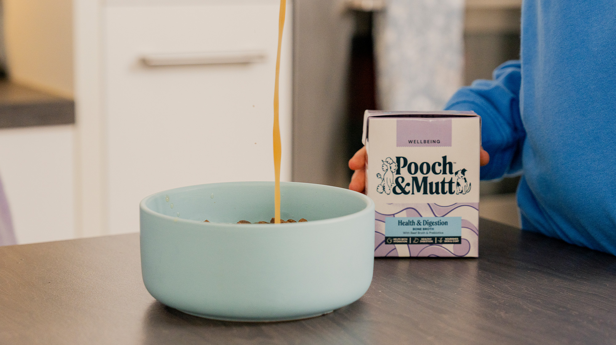 A dog owner pours Pooch & Mutt's Health & Digestion Bone Broth into a bowl of dog food sat on a kitchen counter whilst holding the packaging. 