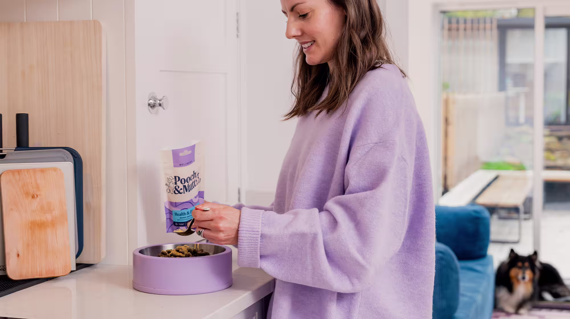 A dog owner preparing some Pooch & Mutt food in a dog bowl whilst their dog lies down in the background.