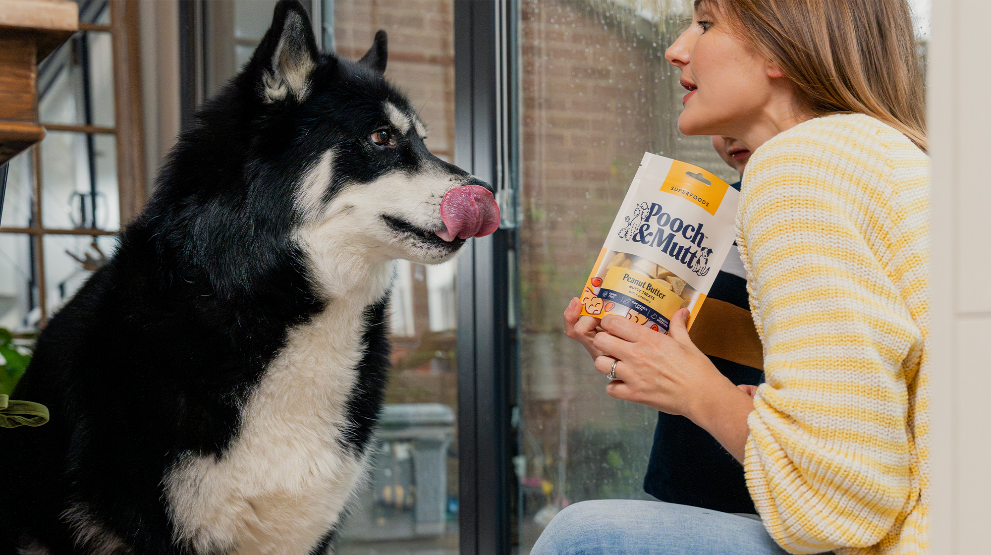 A Siberian husky sits, licking his lips as their own sits in front of them with a bag of Pooch & Mutt Meaty Treats.