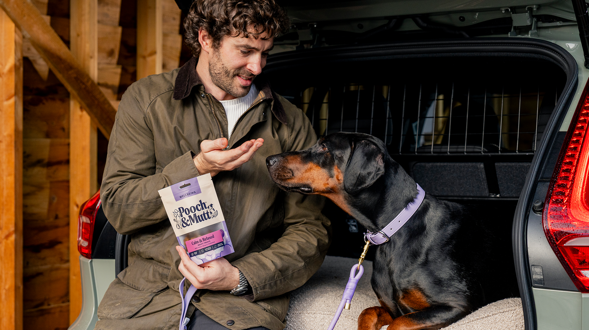 A dog owner sits with their Doberman in the boot of a car whilst they feed them Pooch & Mutt Calm & Relaxed Meaty Treats.