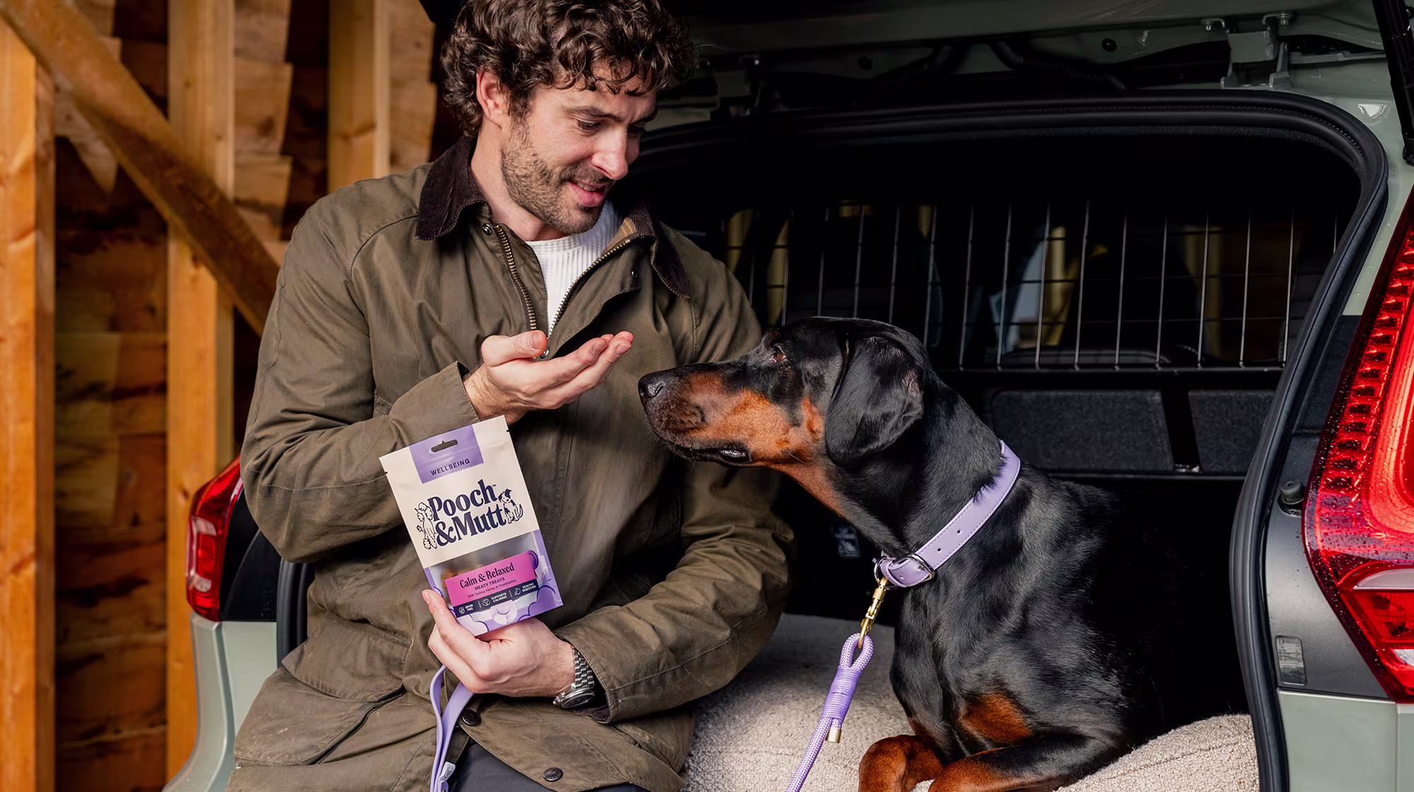A dog owner sits with their Doberman in the boot of a car whilst they feed them Pooch & Mutt Calm & Relaxed Meaty Treats.