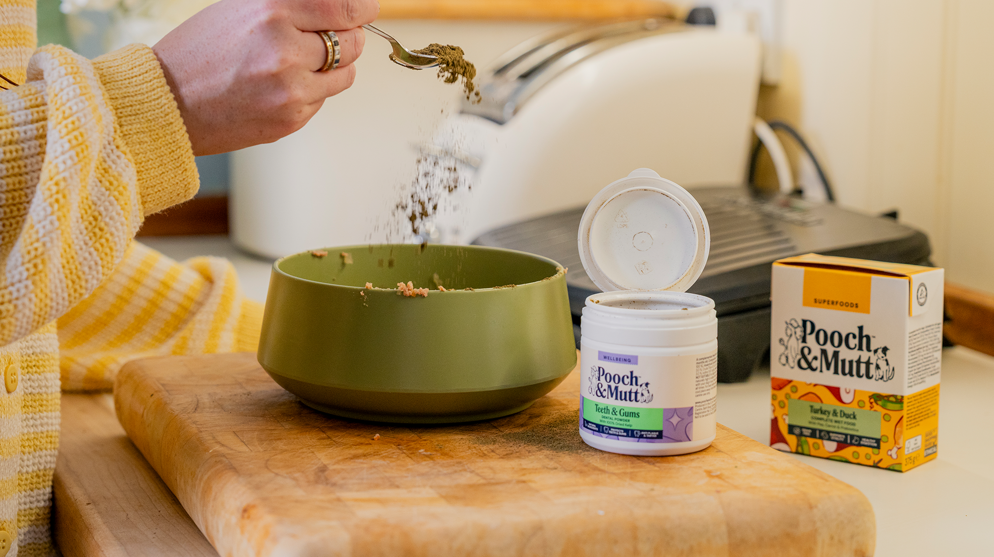 Pooch & Mutt's dental powder being sprinkled from a tea spoon over some dog food in a green bowl, with a pack of Turkey & Duck Wet Food next to it.