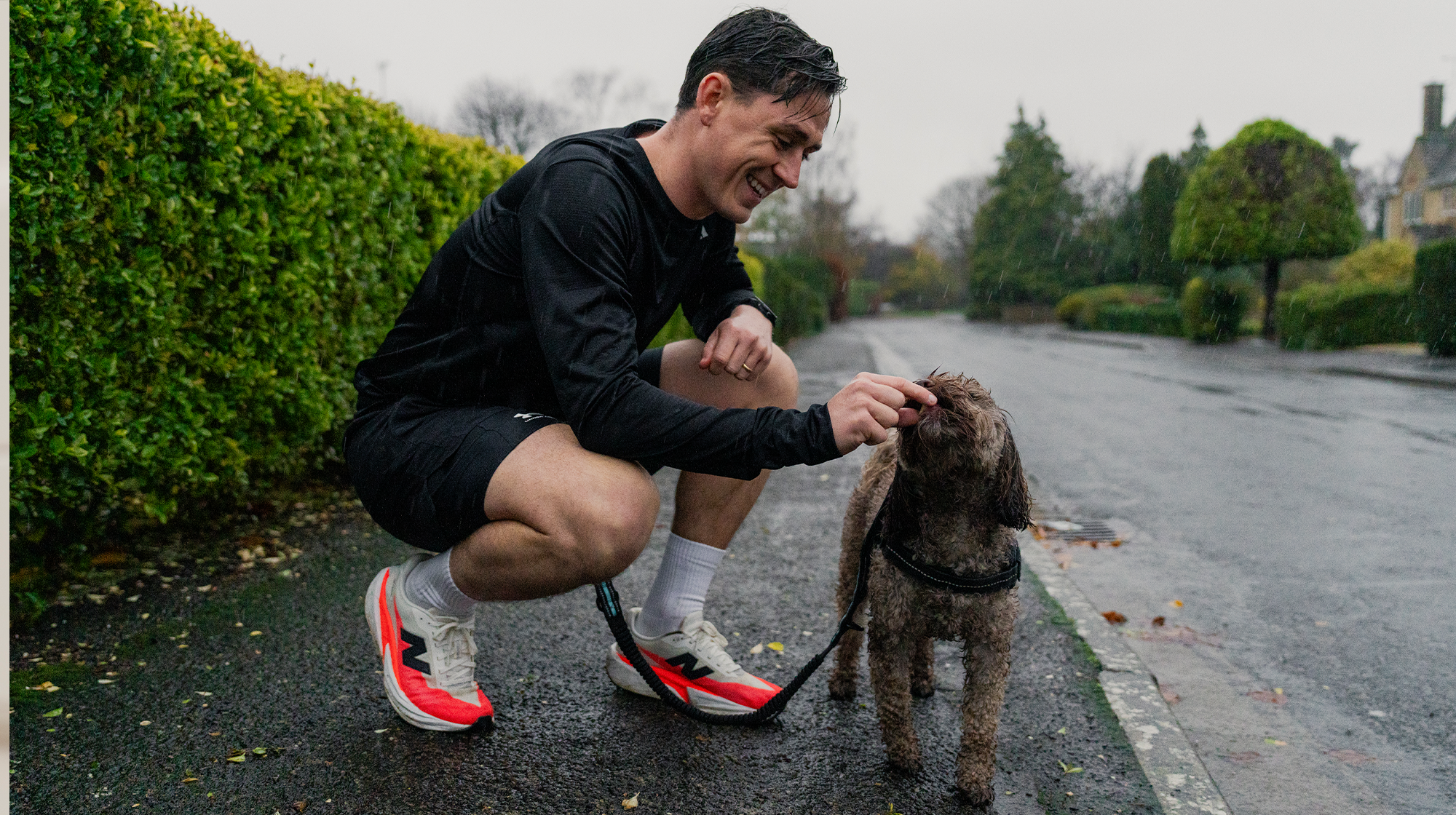 A dog owner crouches down outside whilst wearing running gear, and feeds their brown fluffy dog a treat.