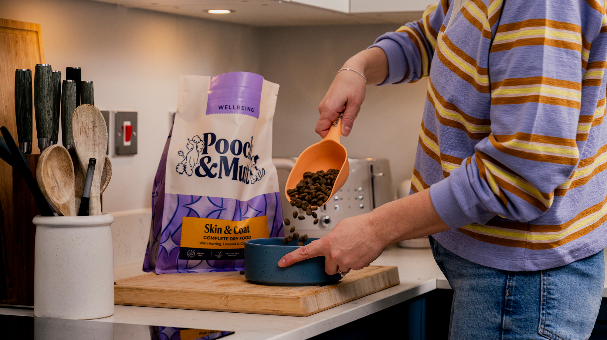 Someone pouring Pooch & Mutt's Skin & Coat Dry Food into a bowl sat on a kitchen counter, with the bag sat behind it.