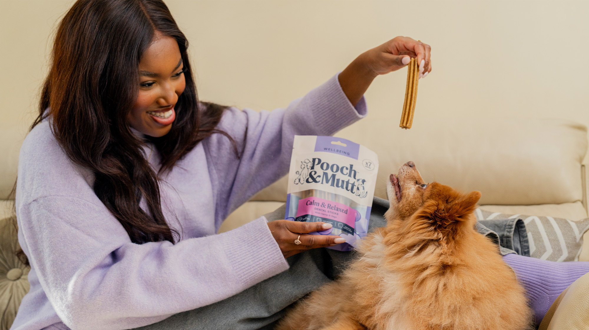 Sat on a sofa, a dog owner dangles a dental chew above dog who is looking up trying to reach it