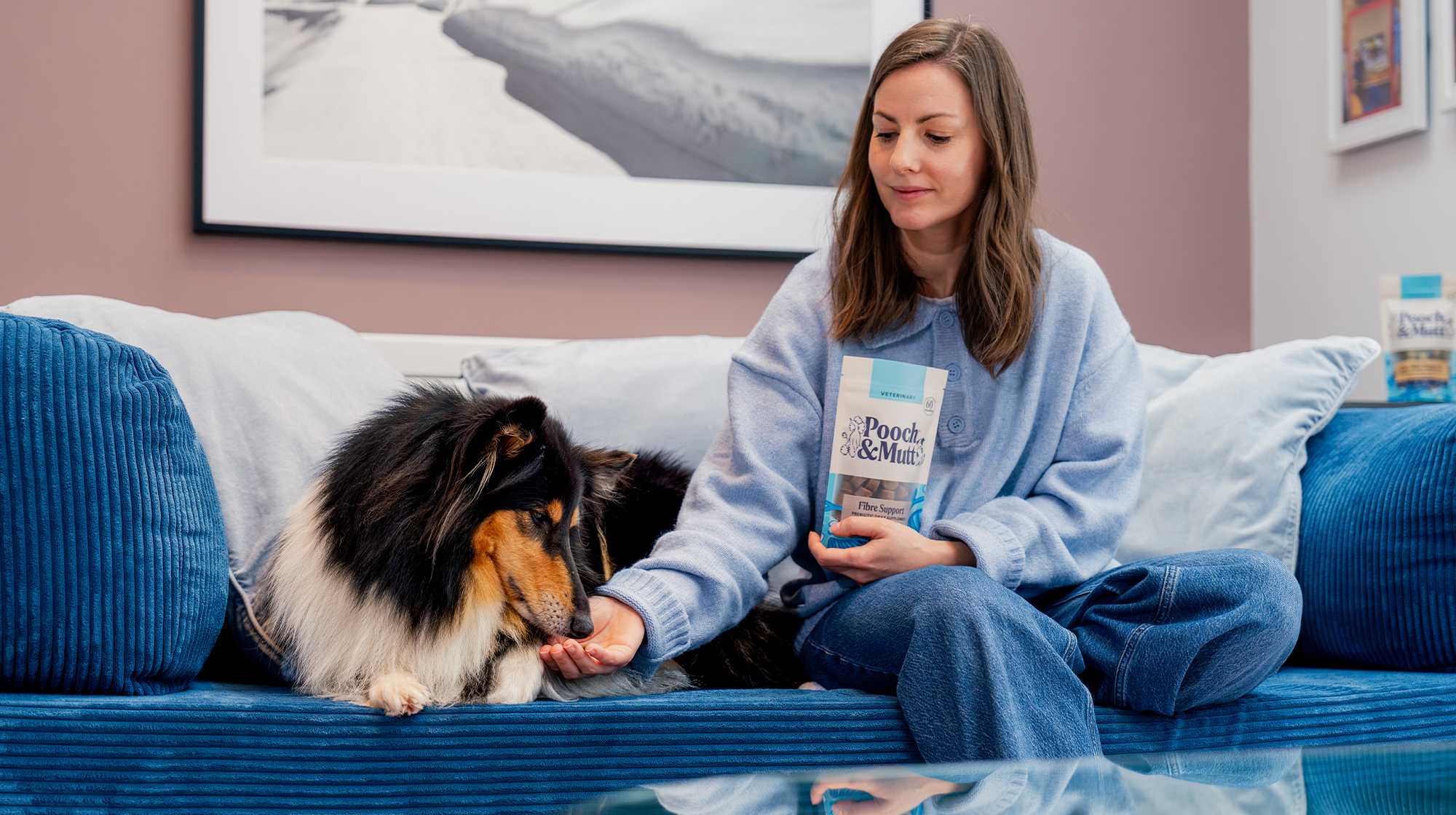 A dog owner feeds their dog from a packet of Pooch & Mutt Meaty Treats whilst sat on the sofa.
