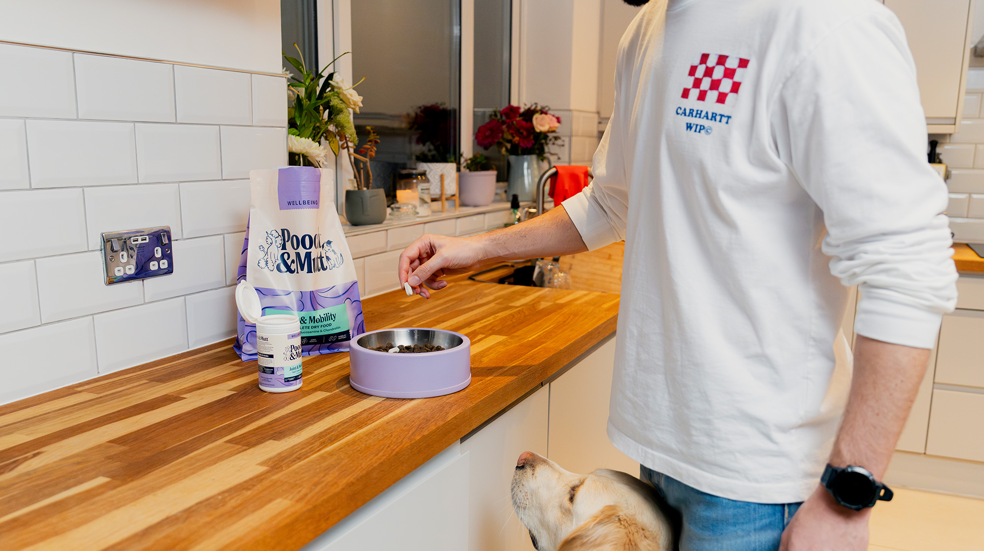 A dog owner in the kitchen preparing a bowl of food using Pooch & Mutt's Joint Care Supplements and Joint Care Dry Food whilst their dog sits at their feet waiting patiently. 
