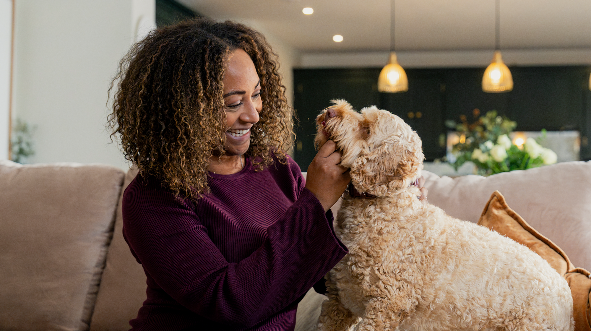 A dog owner in a purple jumper sits on a sofa, fussing their brown, fluffy dog.