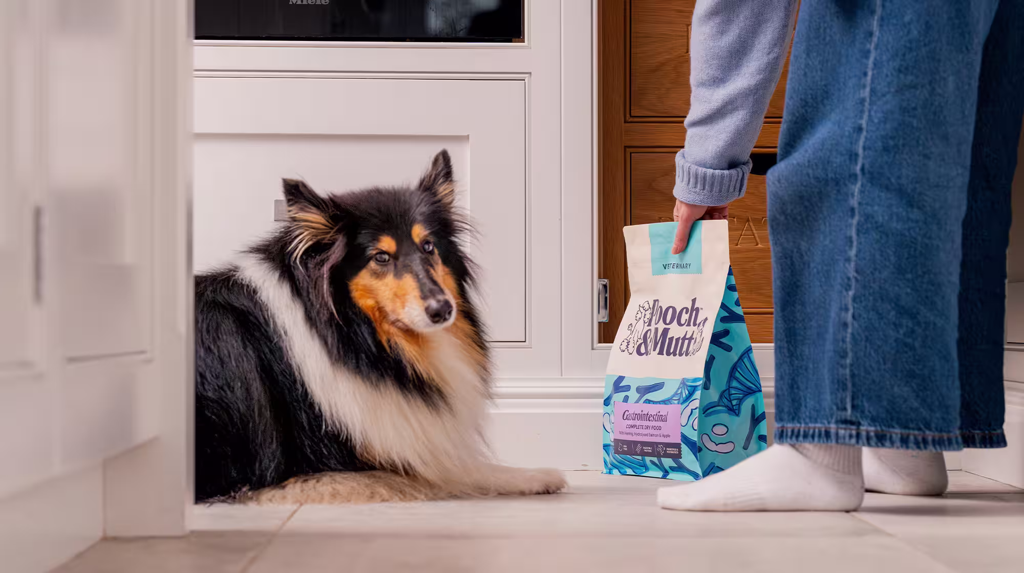 A fluffy dog lays on the floor whilst their owner shows them a bag of Pooch & Mutt's Gastrointestinal Veterinary Dry Food.