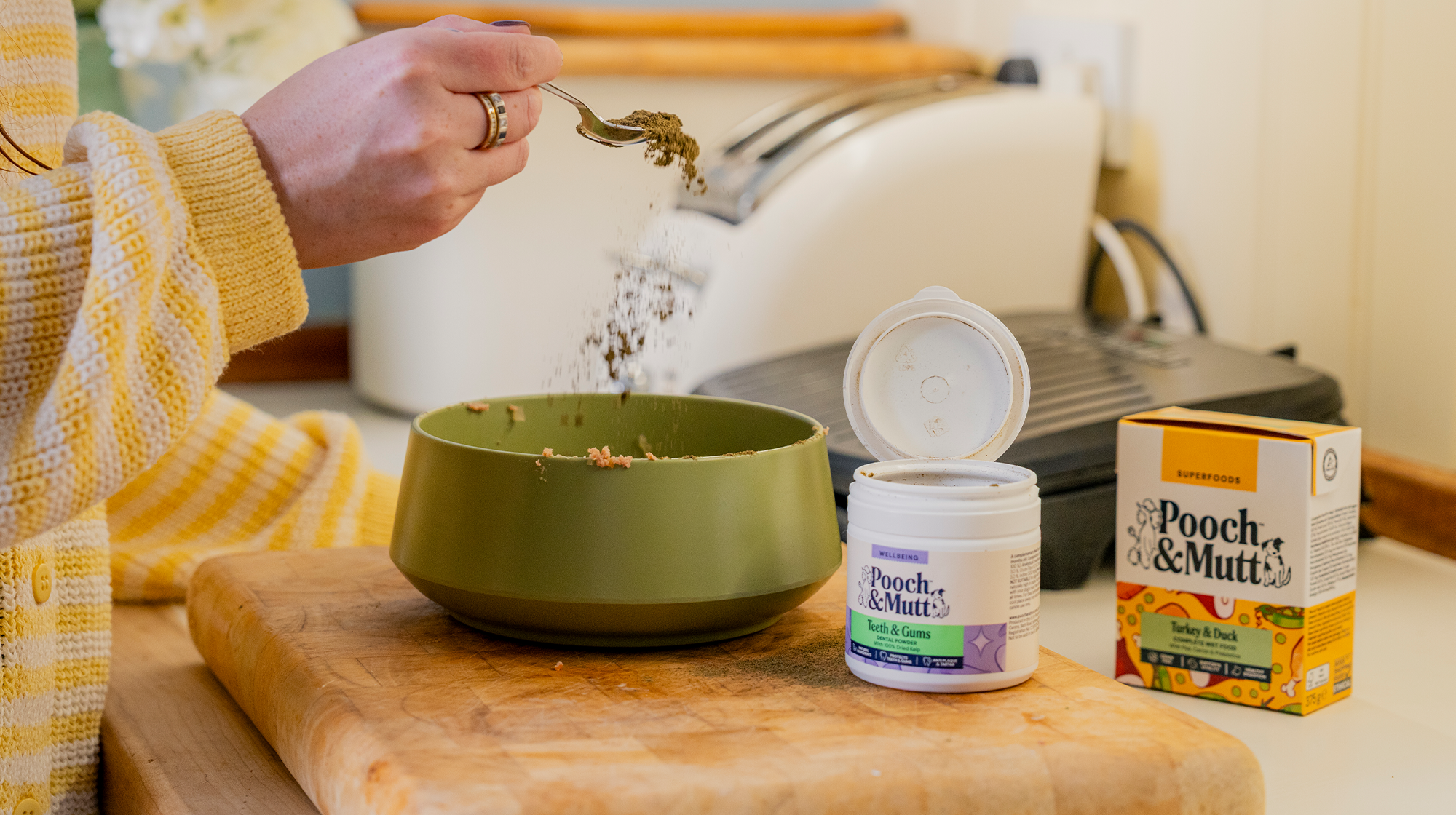 A dog owner sprinkles Pooch & Mutt's Dental Powder over a bowl of dog food, with a pack of Pooch & Mutt's Turkey & Duck Wet Food next to it.